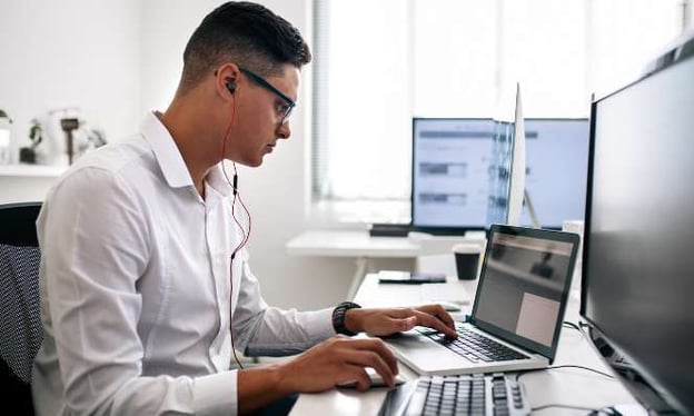 A man with glasses and headphones is working on a laptop beside a desktop monitor at a desk. He is wearing a white dress shirt.